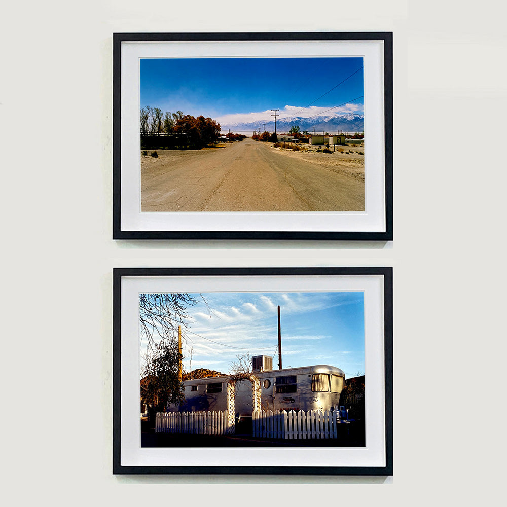 Two black framed photographs by Richard Heeps. The top photograph shows a dusty road in the middle, heading towards the snow capped mountains in the distance, on the right are brown bushes and trees and on the left, single level concrete buildings. The bottom photograph has a rounded metal caravan surrounded by a white picket fence with an archway entrance.