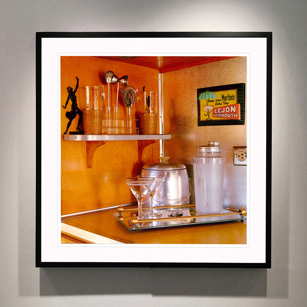 Black framed photograph by Richard Heeps. A drinks corner with martini glasses, cocktail shaker and ice bucket. There is a shelf with cocktail making equipment and it is set against retro orange walls.