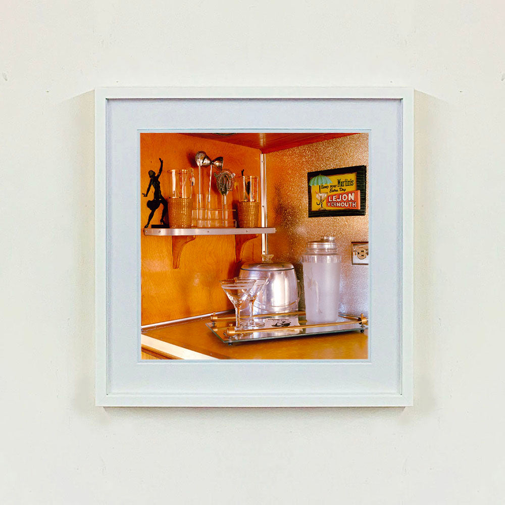 White framed photograph by Richard Heeps. A drinks corner with martini glasses, cocktail shaker and ice bucket. There is a shelf with cocktail making equipment and it is set against retro orange walls.