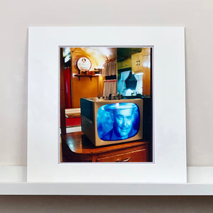 Mounted photograph by Richard Heeps. A retro small brown television sits on a wooden table displaying a black and white film, with the face of Clark Gable filling the screen, it shows as blue and white. In the background of the television is a warm wood-panelled light.