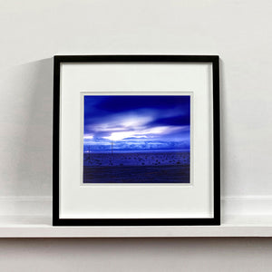 Black framed photograph by Richard Heeps. A blue light hits vast land, mountains and a big sky.