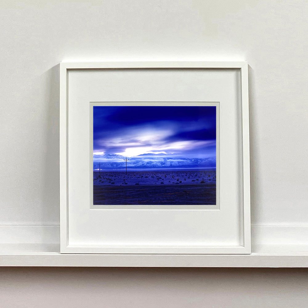 White framed photograph by Richard Heeps. A blue light hits vast land, mountains and a big sky.
