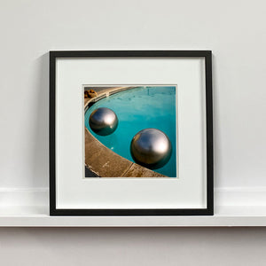 Black framed photograph by Richard Heeps. The corner of a circular swimming pool with two metallic silver beach balls floating on the water.