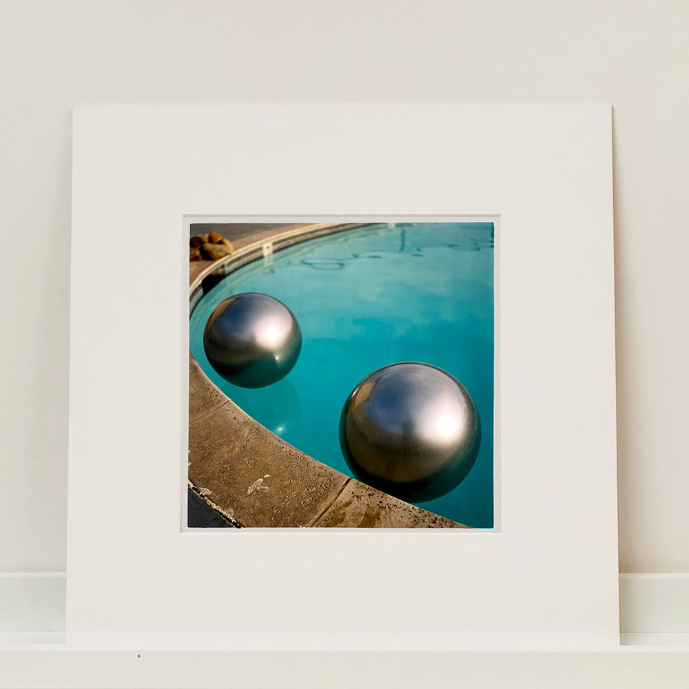 Mounted photograph by Richard Heeps. The corner of a circular swimming pool with two metallic silver beach balls floating on the water.
