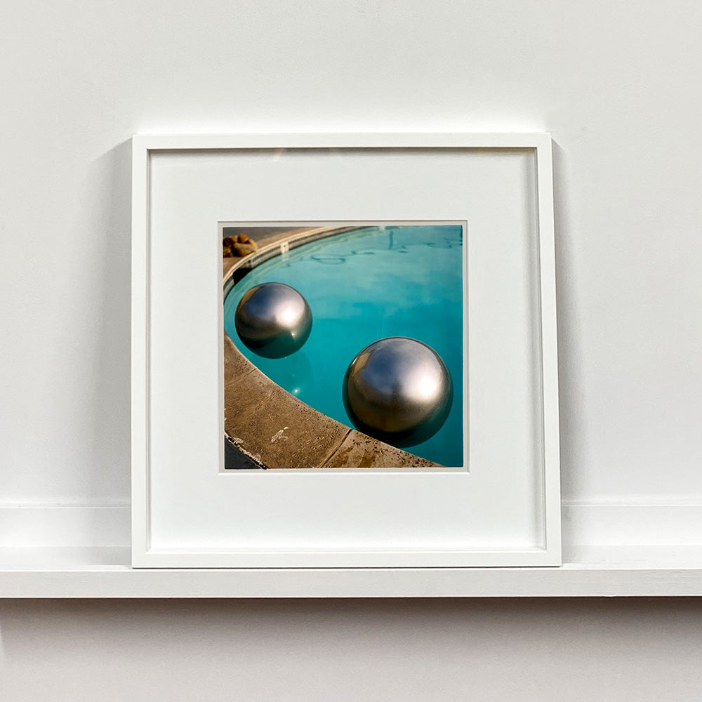 White framed photograph by Richard Heeps. The corner of a circular swimming pool with two metallic silver beach balls floating on the water.