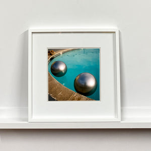 White framed photograph by Richard Heeps. The corner of a circular swimming pool with two metallic silver beach balls floating on the water.