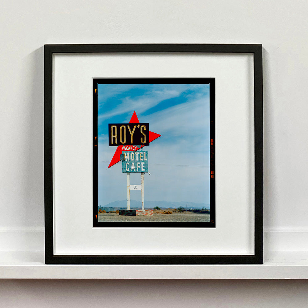 Black framed photograph by Richard Heeps edged by film rebate. A roadside sign on Route 66 in America. The word ROY'S appears in a black sign with a bit red arrow pointing to the left ground, below this VACANCY and on a green square the words MOTEL and CAFE.