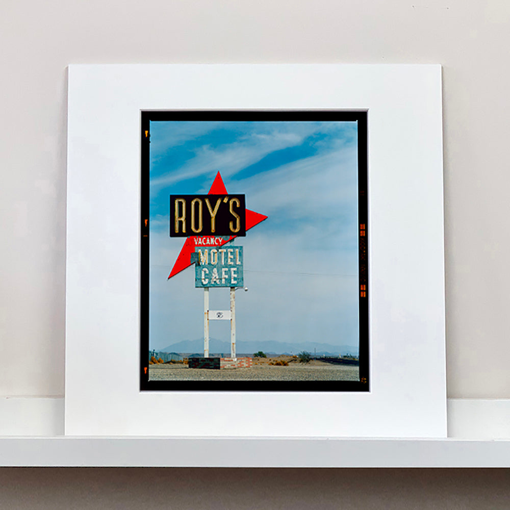 Mounted photograph by Richard Heeps edged by film rebate. A roadside sign on Route 66 in America. The word ROY'S appears in a black sign with a bit red arrow pointing to the left ground, below this VACANCY and on a green square the words MOTEL and CAFE.