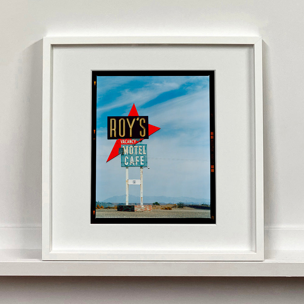 White framed photograph by Richard Heeps edged by film rebate. A roadside sign on Route 66 in America. The word ROY'S appears in a black sign with a bit red arrow pointing to the left ground, below this VACANCY and on a green square the words MOTEL and CAFE.