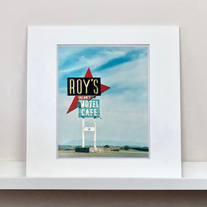 Mounted photograph by Richard Heeps. A roadside sign on Route 66 in America. The word ROY'S appears in a black sign with a big red arrow pointing to the left ground, below this VACANCY and on a green square the words MOTEL and CAFE.