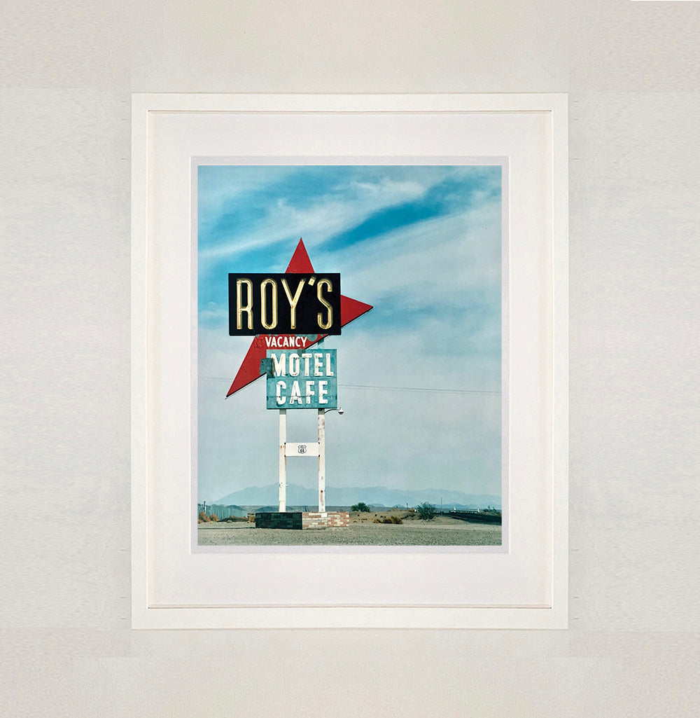 White framed photograph by Richard Heeps. A roadside sign on Route 66 in America. The word ROY'S appears in a black sign with a big red arrow pointing to the left ground, below this VACANCY and on a green square the words MOTEL and CAFE.