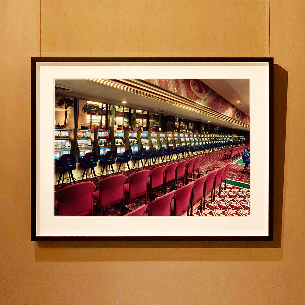 Black framed photograph by Richard Heeps.  The photograph depicts a line of slot machines sitting in a vintage Las Vegas casino. Perspective moves the slot machines from big machines on the left hand side to smaller on the right hand side. In front of the machines are a row of neatly lined blue stools one per machine and then red spectators chairs are neatly lined sitting forefront of the shot.