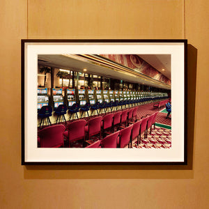 Black framed photograph by Richard Heeps.  The photograph depicts a line of slot machines sitting in a vintage Las Vegas casino. Perspective moves the slot machines from big machines on the left hand side to smaller on the right hand side. In front of the machines are a row of neatly lined blue stools one per machine and then red spectators chairs are neatly lined sitting forefront of the shot.