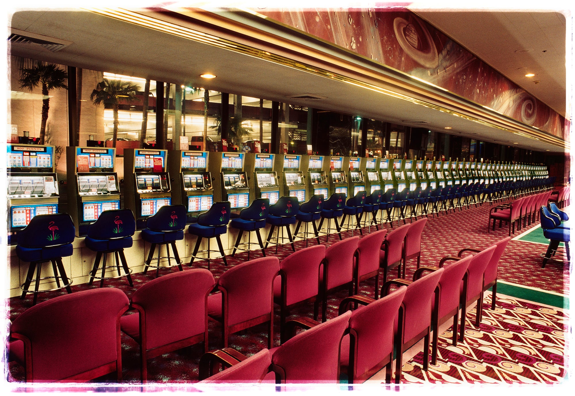 Photograph by Richard Heeps. A line of slot machines sits in a vintage Las Vegas casino. Perspective moves the slot machines from big machines on the left hand side to smaller on the right hand side. In front of the machines are a row of neatly lined blue stools one per machine and then red spectators chairs are neatly lined sitting forefront of the shot.