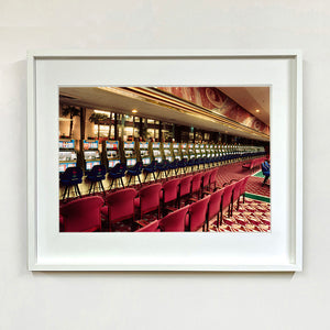 White framed photograph by Richard Heeps. A line of slot machines sits in a vintage Las Vegas casino. Perspective moves the slot machines from big machines on the left hand side to smaller on the right hand side. In front of the machines are a row of neatly lined blue stools one per machine and then red spectators chairs are neatly lined sitting forefront of the shot.