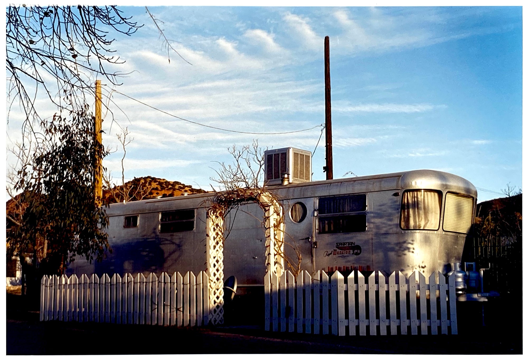 Photograph by Richard Heeps. A vintage American aluminum caravan home surrounded by a white picket fence and at the front a white arch.