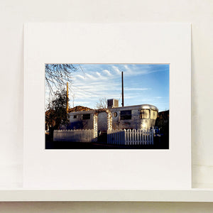 Mounted photograph by Richard Heeps. A vintage American aluminum caravan home surrounded by a white picket fence and at the front a white arch.