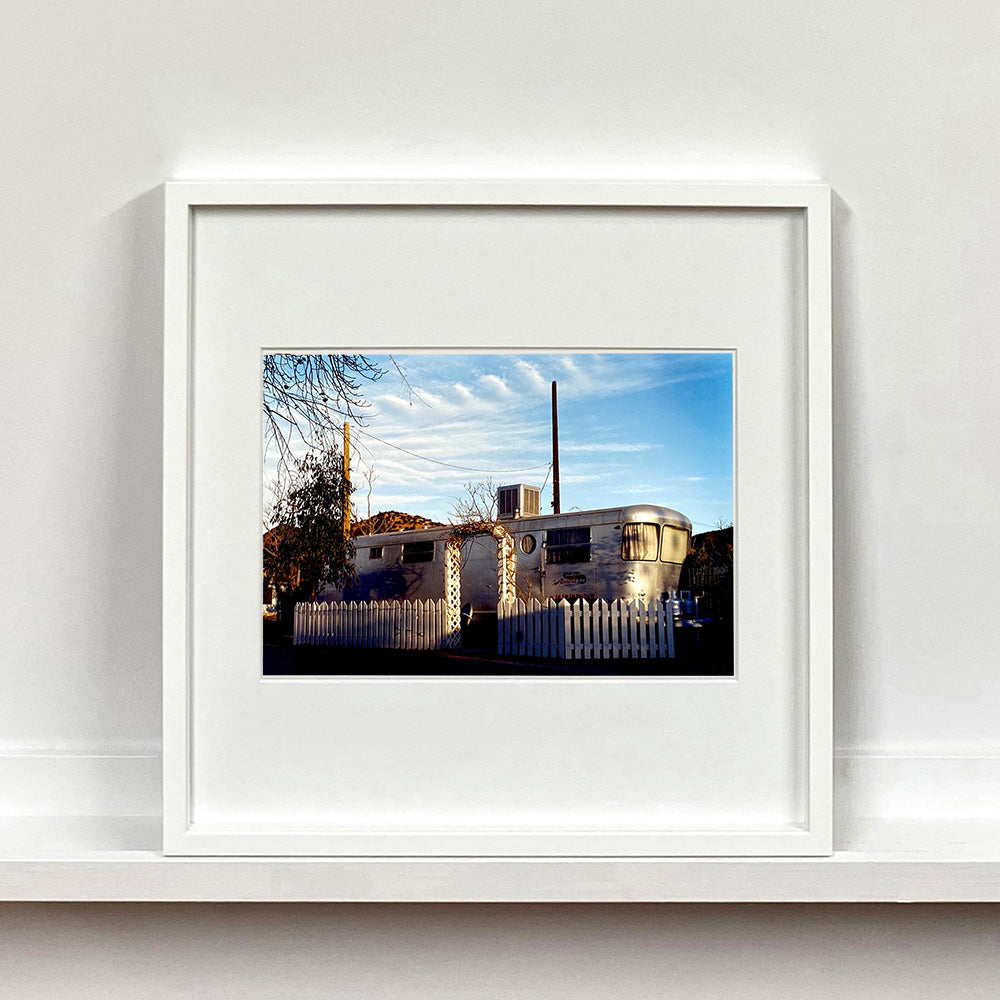 White framed photograph by Richard Heeps. A vintage American aluminum caravan home surrounded by a white picket fence and at the front a white arch.