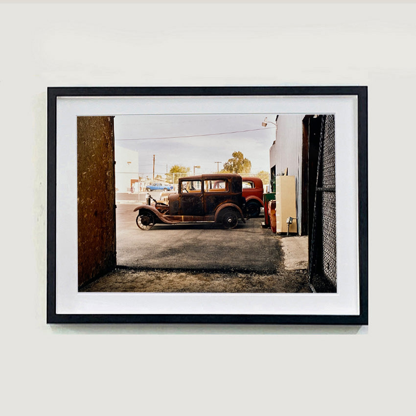 Black framed photograph by Richard Heeps. The side view of two vintage early Ford Motor vehicles parked in a yard. 