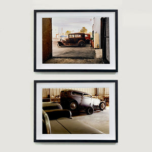 Two black framed photographs by Richard Heeps. The one at the top is of the side view of two vintage early Ford Motor vehicles parked in a yard. The bottom photo is another vintage car in a garage, broken, sitting on a car trailer.