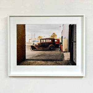 White framed photograph by Richard Heeps. The side view of two vintage early Ford Motor vehicles parked in a yard. 