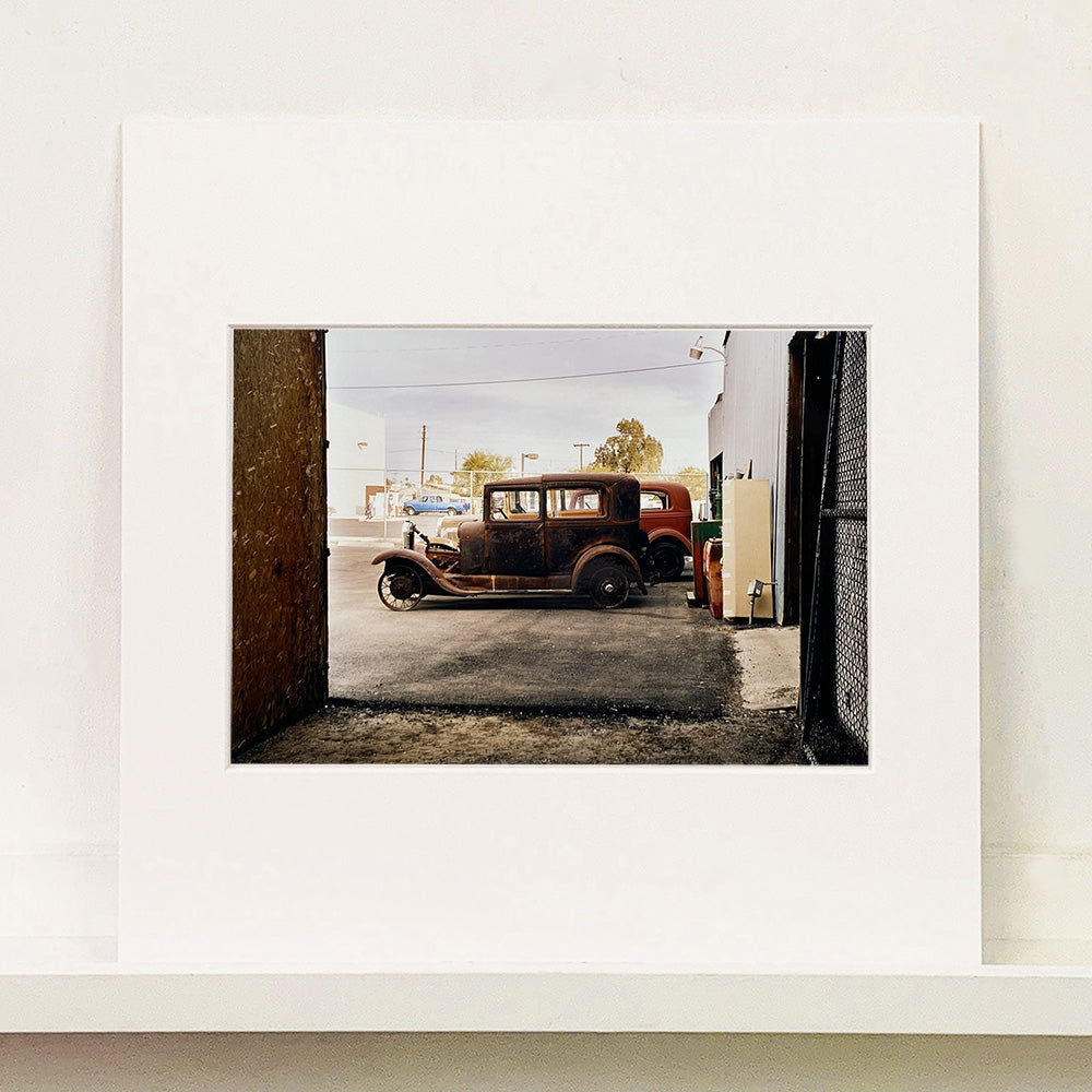 Mounted photograph by Richard Heeps. The side view of two vintage early Ford Motor vehicles parked in a yard. 
