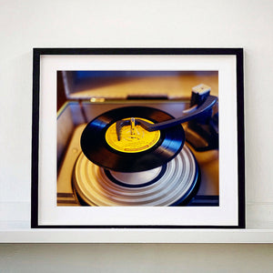 Black frame photograph by Richard Heeps. A black vinyl with a yellow label sits high on the spindle of a deck under the overarm.