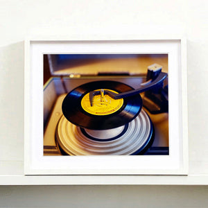 White frame photograph by Richard Heeps. A black vinyl with a yellow label sits high on the spindle of a deck under the overarm.