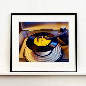 Black frame photograph by Richard Heeps. A black vinyl with a yellow label sits high on the spindle of a deck under the overarm.