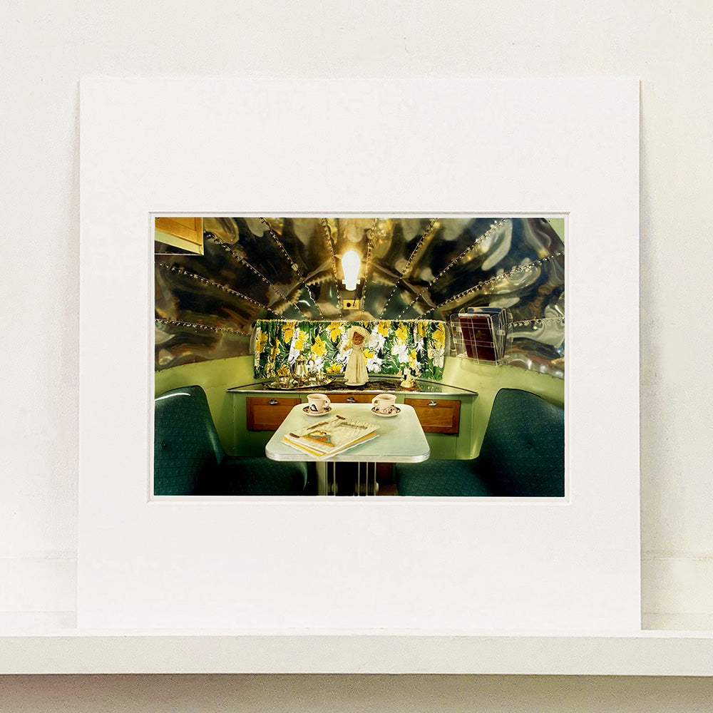 Mounted photograph by Richard Heeps. Inside a trailer, there is a fixed table and chairs with two tea cups and saucers on the table. Behind on the shelf is a tea set and a doll dressed as a bride.