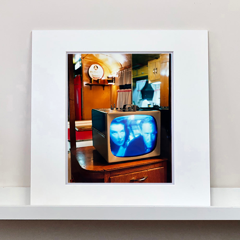 Mounted photograph by Richard Heeps. A retro small brown television sits on a wooden table displaying a scene from a black and white film, Without Love. The image shows as blue and white. In the background of the television is a warm wood-panelled light.