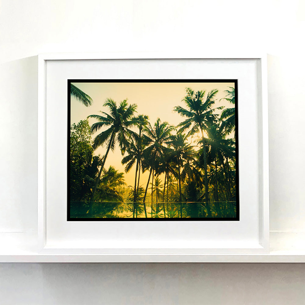 White framed photograph by Richard Heeps. Green palms trees against a warm golden sky, and reflected in a pool below.