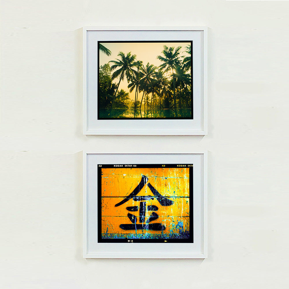 Two white framed photographs by Richard Heeps.  The top photograph is of green palms trees against a warm golden sky, and reflected in a pool below. The bottom photograph is a worn sign, worn out gold background with a Chinese character for gold painted in black.