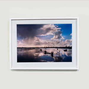 White framed photograph by Richard Heeps. Fenland expanse with water and tufts of grass and an expansive fenland sky, blue with white and grey clouds reflected in the water below.