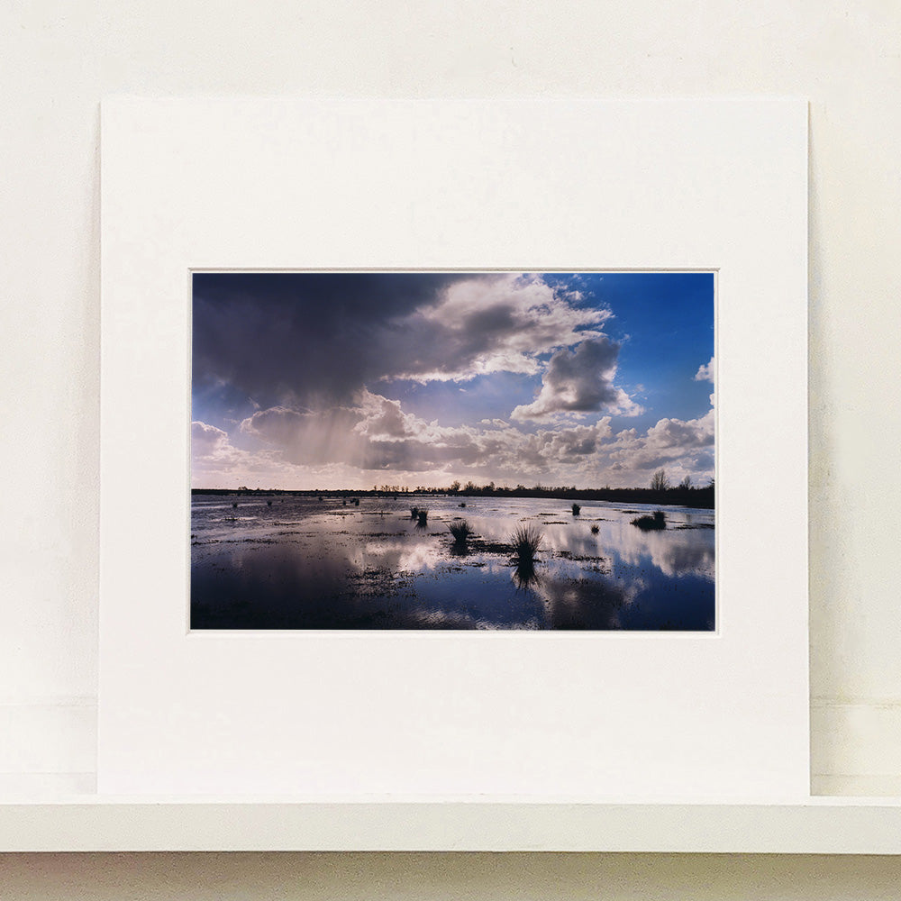Mounted photograph by Richard Heeps. Fenland expanse with water and tufts of grass and an expansive fenland sky, blue with white and grey clouds reflected in the water below.