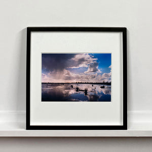 Black framed photograph by Richard Heeps. Fenland expanse with water and tufts of grass and an expansive fenland sky, blue with white and grey clouds reflected in the water below.