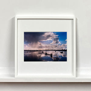 White framed photograph by Richard Heeps. Fenland expanse with water and tufts of grass and an expansive fenland sky, blue with white and grey clouds reflected in the water below.