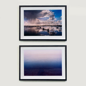 Two black framed photographs by Richard Heeps. The top one is of a Fenland expanse with water and tufts of grass and an expansive fenland sky, blue with white and grey clouds reflected in the water below. The photograph at the bottom is fenland blue water at the bottom fading into one tone white cloud filled sky at the top.