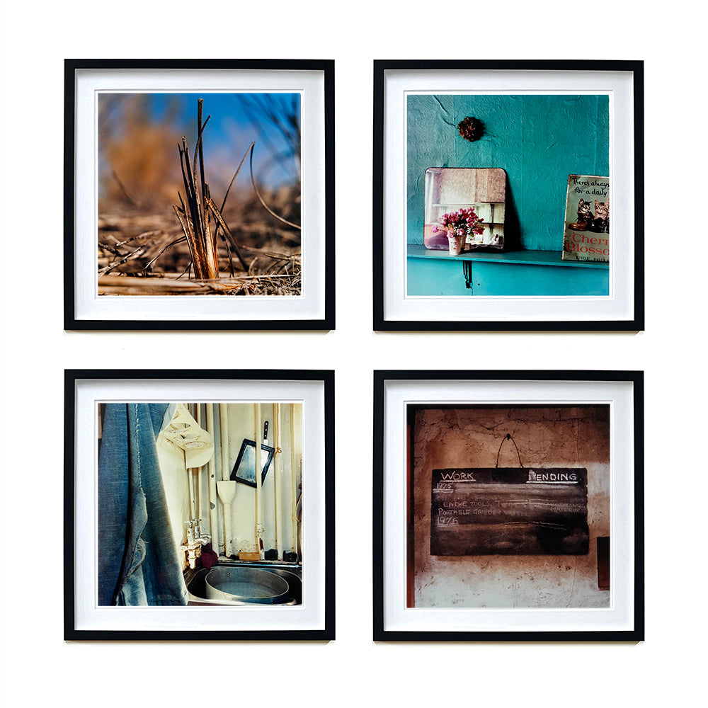 Four black framed photographs in a square by Richard Heeps. The top photograph is of a reed tuft sticking out of a blurred reed bed. The next photograph is a blue shelf, with blue wrinkled wallpaper behind. On the shelf sits a small posy, mirror and cat magazine. The third photograph is a old and worn sink and taps. The fourth photograph is a chalk board with Work Pending chalked on top and the writing beneath rubbed out.