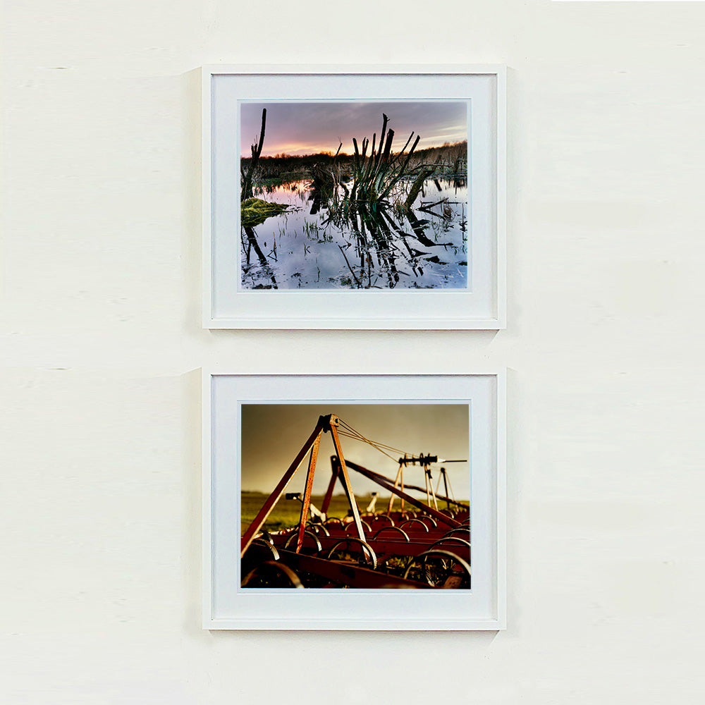 White framed photograph by Richard Heeps. Photograph of cut down, lichen clad branches poking out of the flooded fen field. The branches are strikingly dark and create dark reflections with a golden sunset in the background. The bottom photograph is of the wheels of an orangy/rusty looking plough bathed in the evening dusk light.