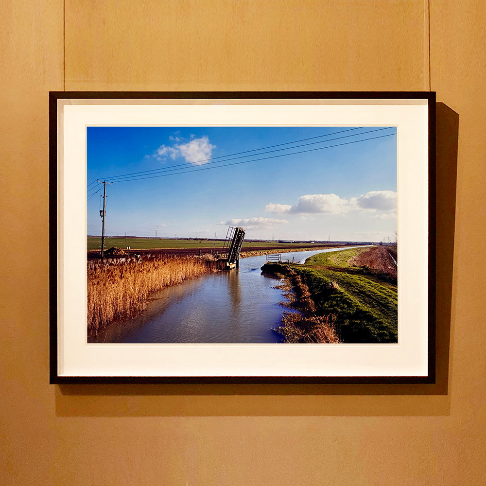 Black framed photograph by Richard Heeps. Fenland waterway with a weight drain up in the middle. Fenland sits either side and there is a blue sky.