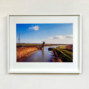 White framed photograph by Richard Heeps. Fenland waterway with a weight drain up in the middle. Fenland sits either side and there is a blue sky.