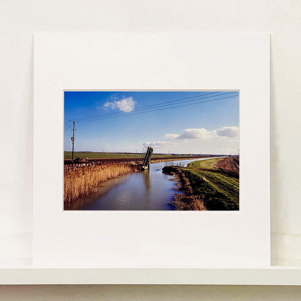Photograph by Richard Heeps. Fenland waterway with a weight drain up in the middle. Fenland sits either side and there is a blue sky.
