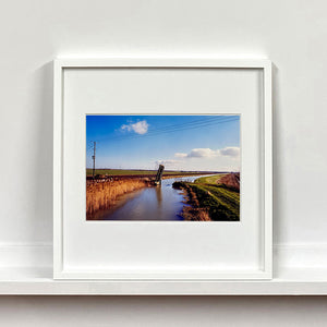 White framed photograph by Richard Heeps. Fenland waterway with a weight drain up in the middle. Fenland sits either side and there is a blue sky.