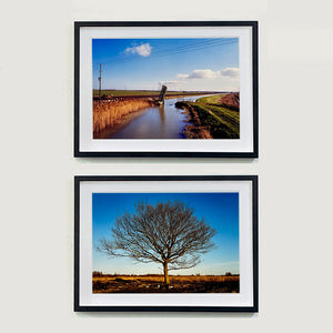Two black framed photographs by photographer Richard Heeps. The top one is of a fenland waterway with a weight drain up in the middle. Fenland sits either side and there is a blue sky. The bottom photo is a bare winter tree against a blue sky.