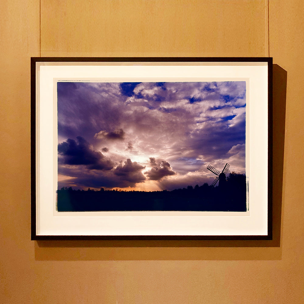 Black framed photograph by Richard Heeps. A silhouette of a tree lined fen flat land with a windpump sitting on the right hand side. The sky is cloudy and bathed in golden dusk light.