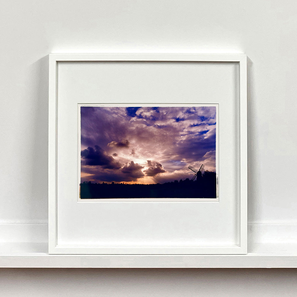 White framed photograph by Richard Heeps. A silhouette of a tree lined fen flat land with a windpump sitting on the right hand side. The sky is cloudy and bathed in golden dusk light.