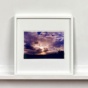 White framed photograph by Richard Heeps. A silhouette of a tree lined fen flat land with a windpump sitting on the right hand side. The sky is cloudy and bathed in golden dusk light.