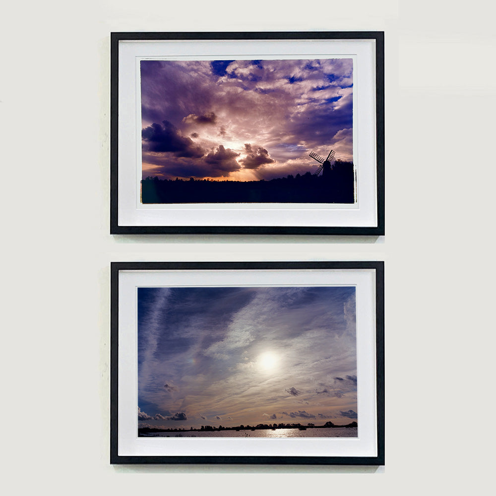 Two black framed photographs by Richard Heeps. The top one is of a silhouette of a tree lined fen flat land with a windpump sitting on the right hand side. The sky is cloudy and bathed in golden dusk light. The bottom photograph has the flat fen land with a stretch of water in front of it. This is covered by a vast fen dusk sky.