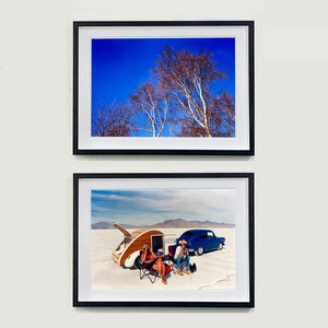 Two black framed photographs by Richard Heeps. The top photograph is looking up at the tops of four leafless silver birches against a deep blue autumn sky. The bottom photograph is a white desert with mountains in the back. At the centre of the photograph is a bright blue car with a small pod caravan behind and in front two women sitting in their camping chairs.